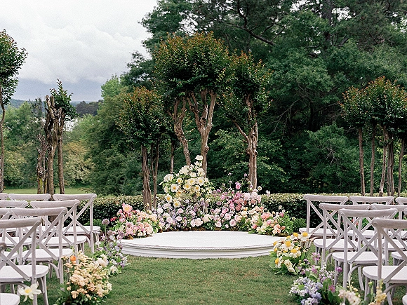 Outdoor ceremony setup with white folding chairs around a round platform, plus low floral aisle arrangements on a lawn under cloudy sky
