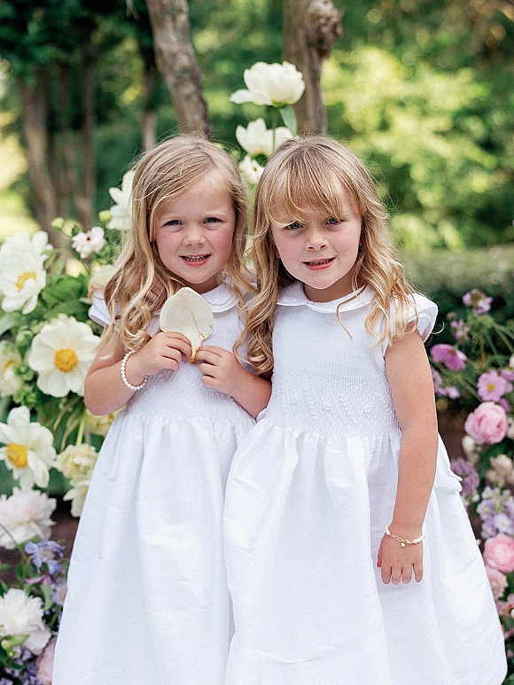 Flower girl portrait in a white flower girl dress holding petals, standing in lush garden greenery with colorful blooms in natural light