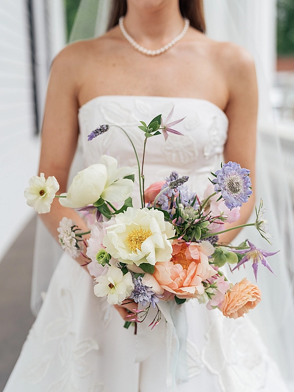 Bridal bouquet with garden style blooms of white flowers, peach roses, purple accents and greenery, held by bride in strapless dress on walkway