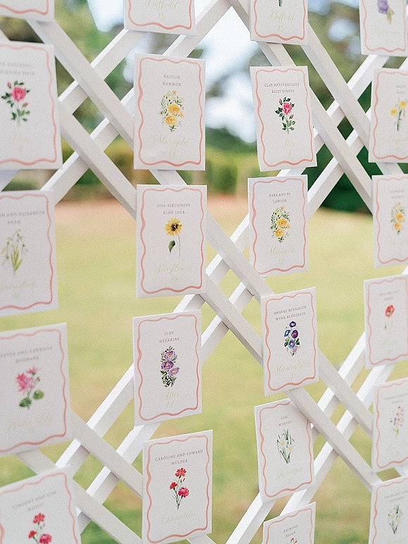 Wedding escort cards arranged on a white lattice escort card display with floral illustrations and calligraphy, set on an outdoor lawn among trees
