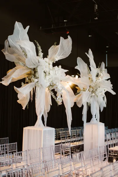 Reception centerpiece with tall wedding centerpieces of white orchids, roses, and baby's breath on pedestal stands before black drape backdrop