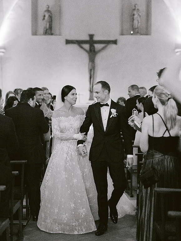 Wedding recessional as bride in long-sleeve lace gown walks arm-in-arm with groom in tuxedo down church aisle past pews and guests
