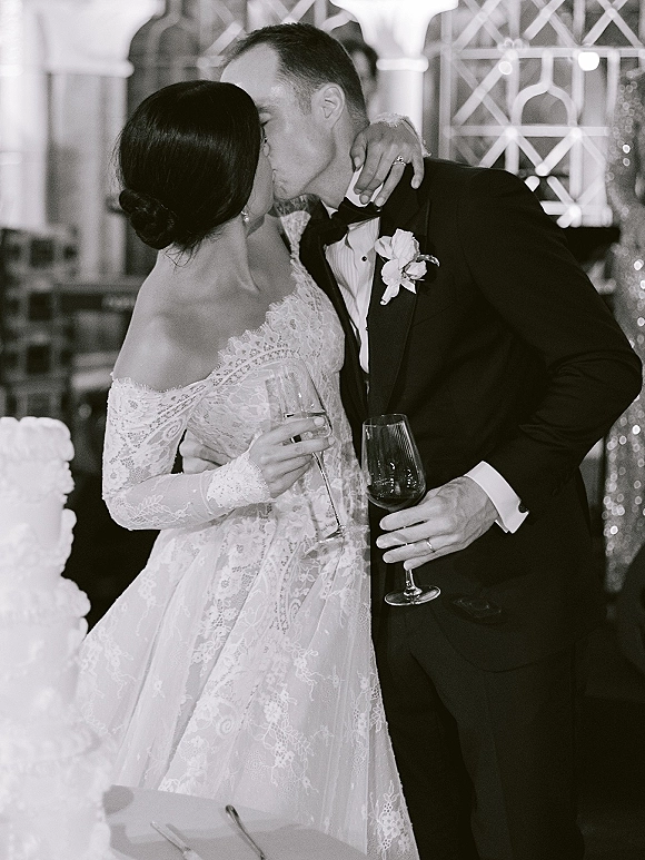 Wedding kiss as the bride and groom kissing while holding champagne flutes beside the wedding cake at a softly lit indoor reception