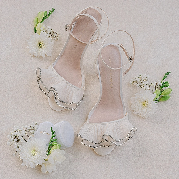 Bridal shoes with rhinestone embellishments and ankle straps in a wedding heels flat lay, styled with white florals and ring box on cream fabric