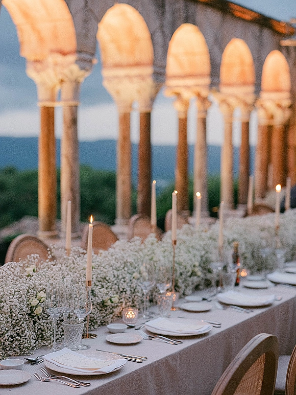 Reception tablescape with long banquet table setting, baby's breath garland, taper candles and glassware on a stone-arched terrace with hills beyond
