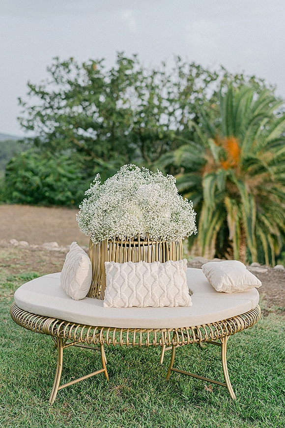 Wedding lounge seating with a round rattan daybed, cream cushions and baby's breath florals set on a grassy lawn with palms and hills