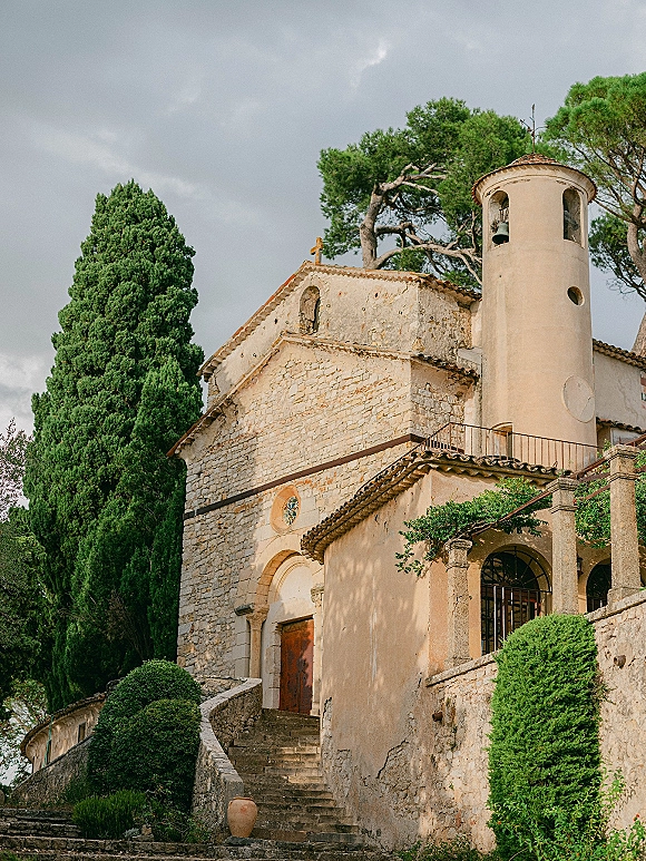 Wedding venue exterior with stone chapel entrance, arched wooden door, bell tower, and stone stairs framed by cypress trees under cloudy sky