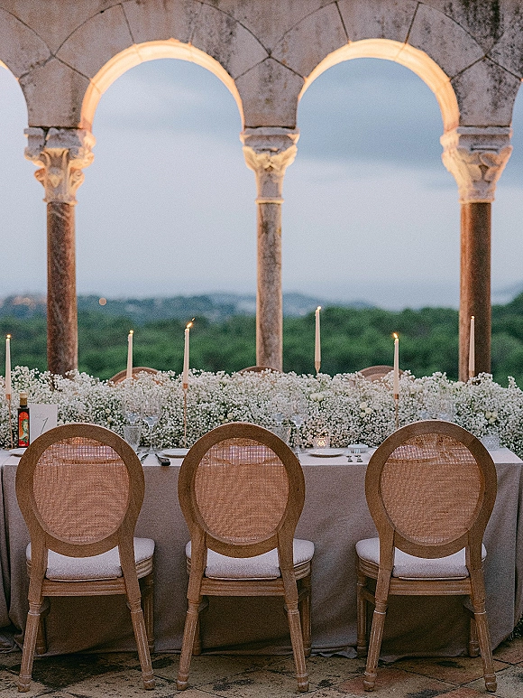 Reception tablescape with head table decor featuring a long beige banquet table, rattan chairs, taper candles, and white floral runner under stone arches