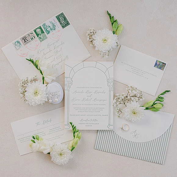 Wedding invitation suite flat lay with mint green cards, calligraphy envelopes, vintage stamps, pearl earrings, and band on neutral fabric
