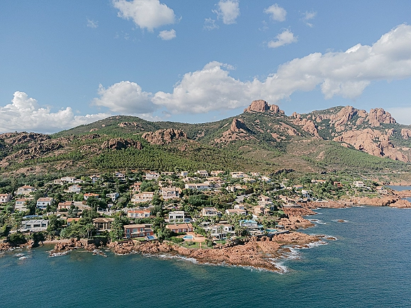 Coastal landscape with rocky coastline aerial view of turquoise ocean, hillside village homes, and distant mountains under blue sky and clouds