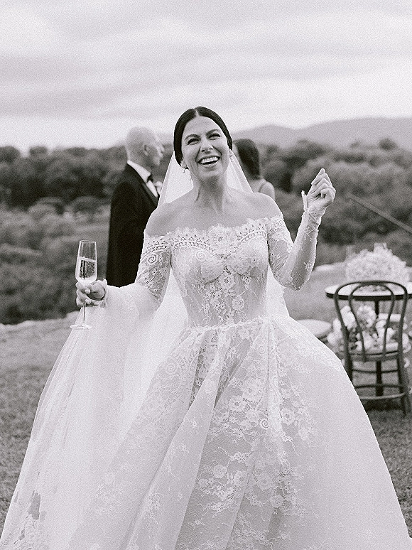 Bride portrait in a black and white wedding portrait, laughing in a lace dress holding a champagne flute at outdoor cocktail hour with hills behind
