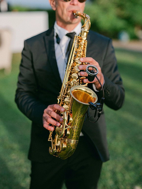 Wedding saxophonist performing live wedding music in a black tuxedo and sunglasses, playing saxophone on an outdoor lawn with greenery