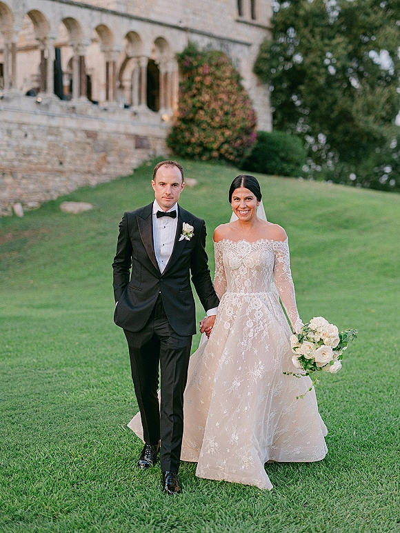 Couple portrait of bride and groom holding hands, with veil and bouquet, on a lawn before a stone building with arched windows