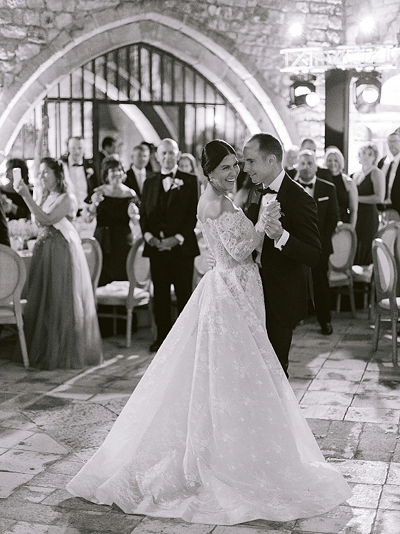First dance as bride in lace wedding dress twirls with groom in tuxedo on courtyard dance floor, guests watching by stone arch and lights