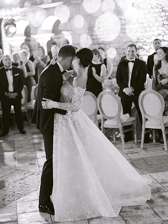 Wedding first dance as bride in a lace long sleeve gown kisses her tuxedo groom under string lights in a stone courtyard with guests seated nearby