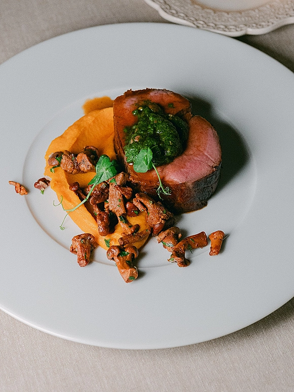 Wedding plated dinner featuring roasted meat with green sauce, vegetable puree, mushrooms and microgreens on a white plate atop linen tablecloth