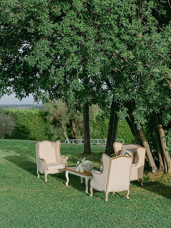 Outdoor lounge seating with cream upholstered armchairs around a coffee table and baby's breath centerpiece on a shaded lawn with trees