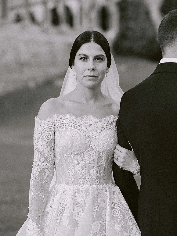 Couple portrait of bride in off the shoulder lace wedding dress holding groom’s arm, her veil flowing on a lawn with stone building behind