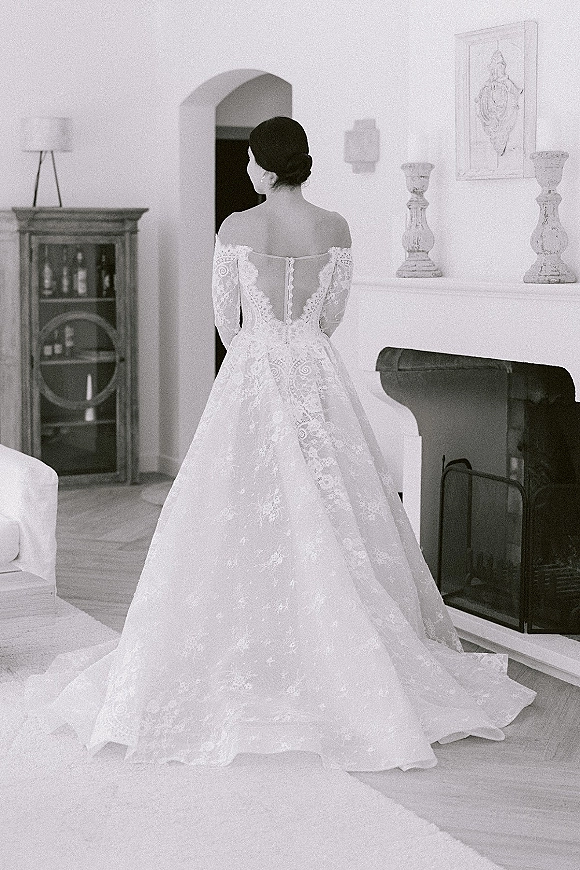 Wedding dress with lace wedding dress details, off-the-shoulder long sleeves and buttoned illusion back, in a living room by the fireplace