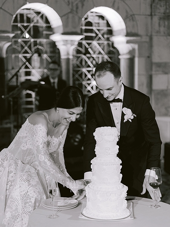 Wedding cake cutting as bride in lace sleeves and groom in tuxedo share the first slice at a reception table by arched windows