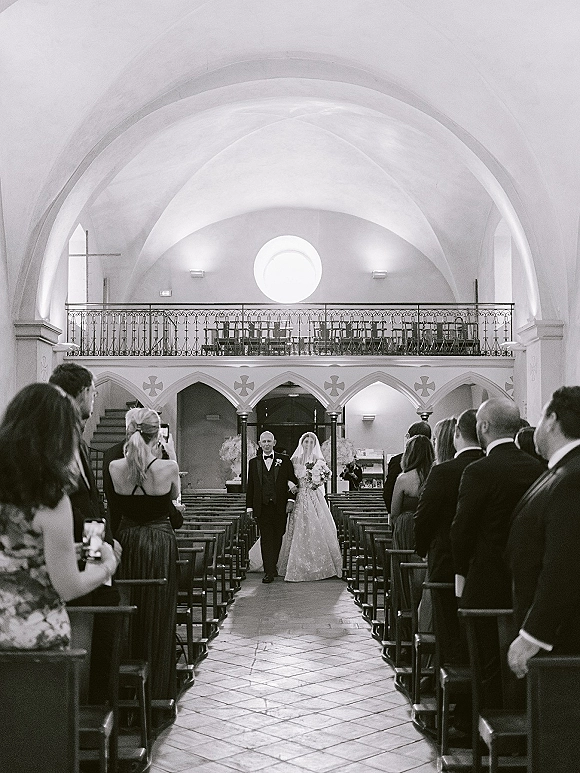 Wedding processional as bride walking down aisle with veil and bouquet, escorted past church pews beneath arched ceiling and stained glass window