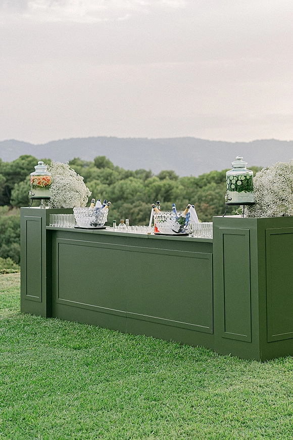Wedding bar setup with glass dispensers of fruit-infused and cucumber water, champagne on ice, and baby’s breath on a mountain lawn