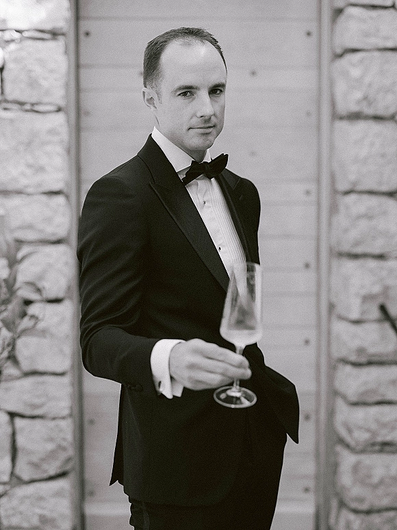 Groom portrait in a black tuxedo, bow tie and cufflinks, holding a champagne flute while leaning by a stone wall and wooden door