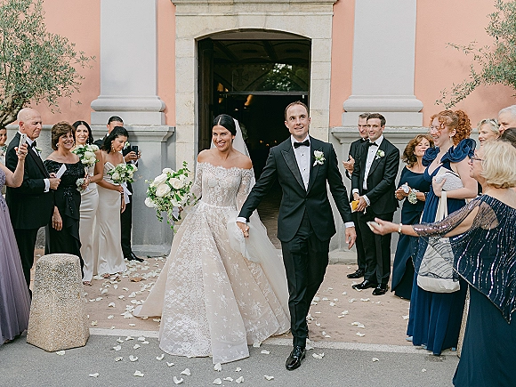 Wedding recessional as bride and groom walk out holding hands, bride with bouquet and veil, guests toss rose petals at church entrance