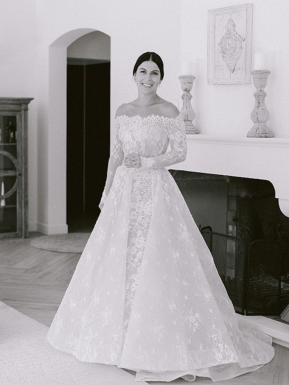 Bridal portrait of a bride in a lace ball gown wedding dress with off-the-shoulder sleeves, posed by an indoor fireplace mantel and art