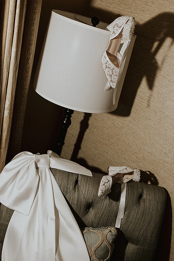 Wedding shoes, lace wedding heels with pearl strap and satin bow styled by a sofa under window light against a textured wall
