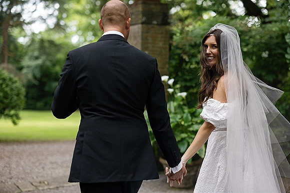Couple portrait of bride and groom holding hands, walking down a garden path as she looks back, long veil trailing near a brick pillar