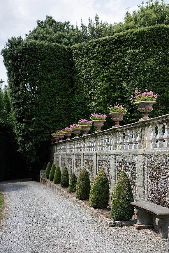 Garden walkway with a stone balustrade and pink flower planters lining a gravel path, framed by trimmed topiary and tall hedges