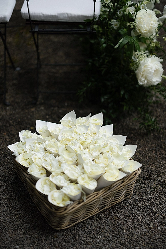 Wedding flower petal cones for a flower petal toss in a wicker basket with white petals and greenery beside a bench on gravel ground