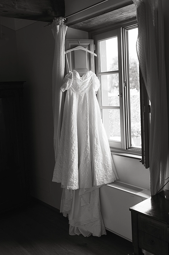 Wedding dress with lace wedding dress details and puff sleeves hanging by a window, framed by soft curtains in an indoor room with beams