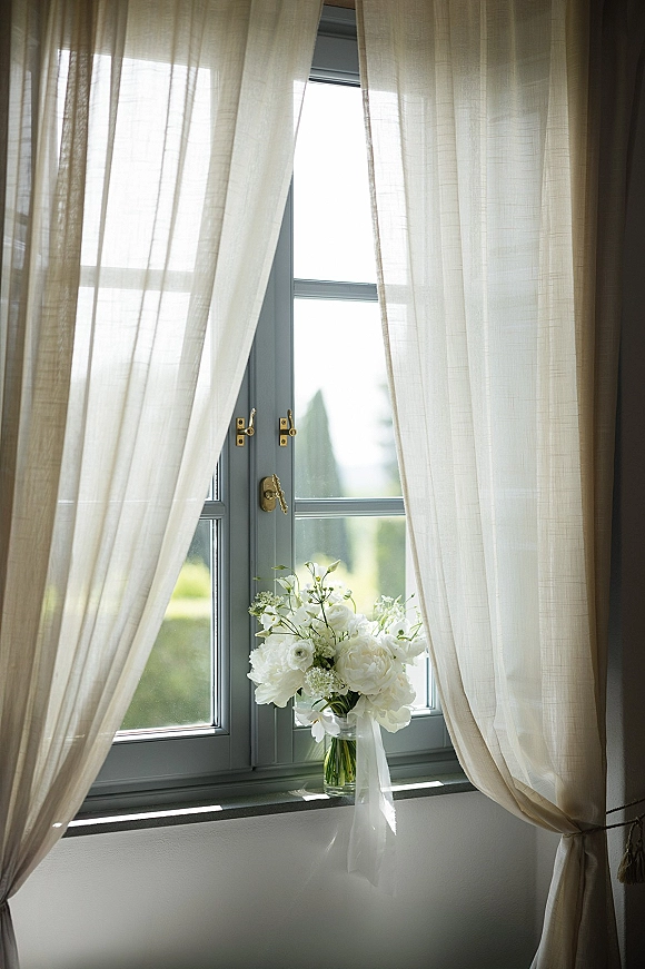 Bridal bouquet of white flowers in a clear glass vase with sheer ribbon on a windowsill, softly lit by window light and greenery outside
