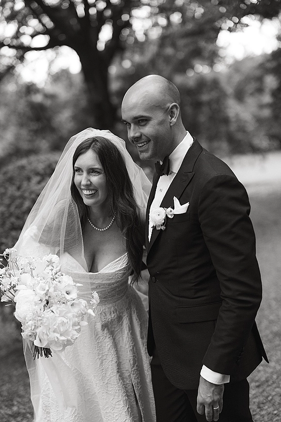 Couple portrait in a black and white wedding portrait style, bride in veil holding bouquet beside groom in tuxedo on a garden lawn