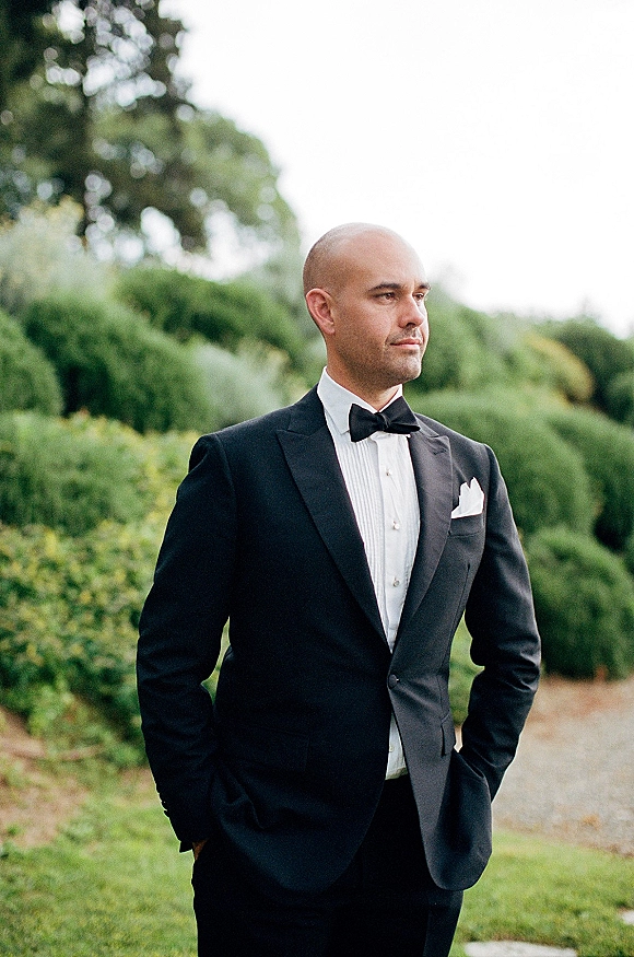 Groom portrait in a classic black tuxedo with bow tie and pocket square, looking away on a gravel path by green hedges