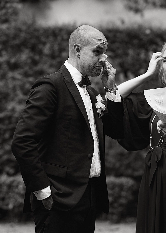 Groom portrait captures him wiping tears in a black tuxedo with bow tie and white rose boutonniere against lush garden greenery