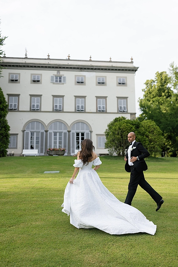 Couple portrait of bride and groom walking on a manicured lawn before a grand villa with arched windows, her long train flowing