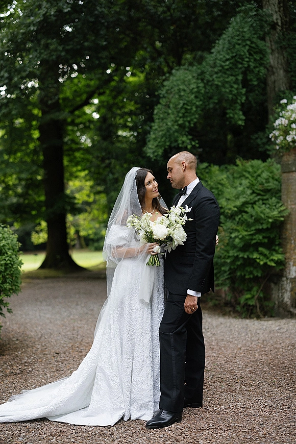 Couple portrait of bride and groom holding hands, bride with white bouquet and long veil, groom in black tuxedo on garden path