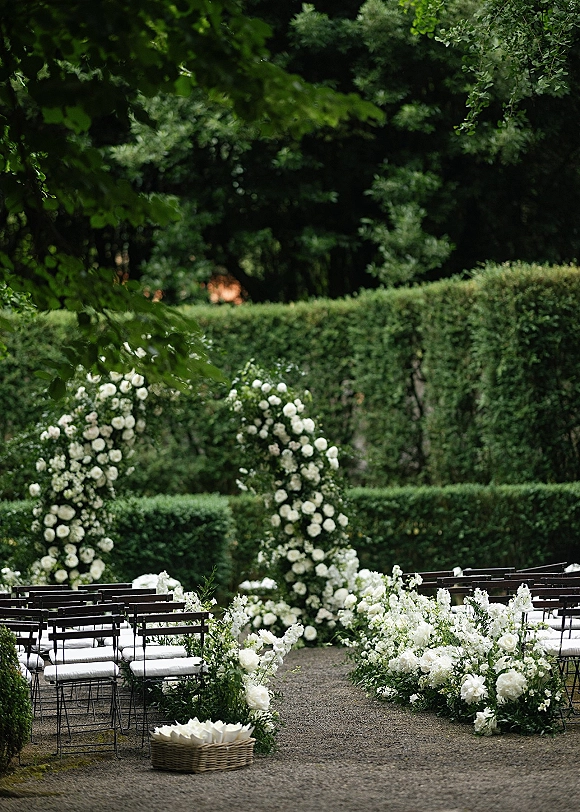 Ceremony aisle decor with grounded white florals leading to a white rose arch, lined by black chairs with cushions in a hedge garden setting