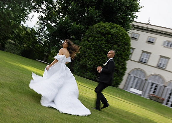 Couple portrait of bride twirling dress as groom chases her, gown train flowing on green lawn beside a manor house with arched windows