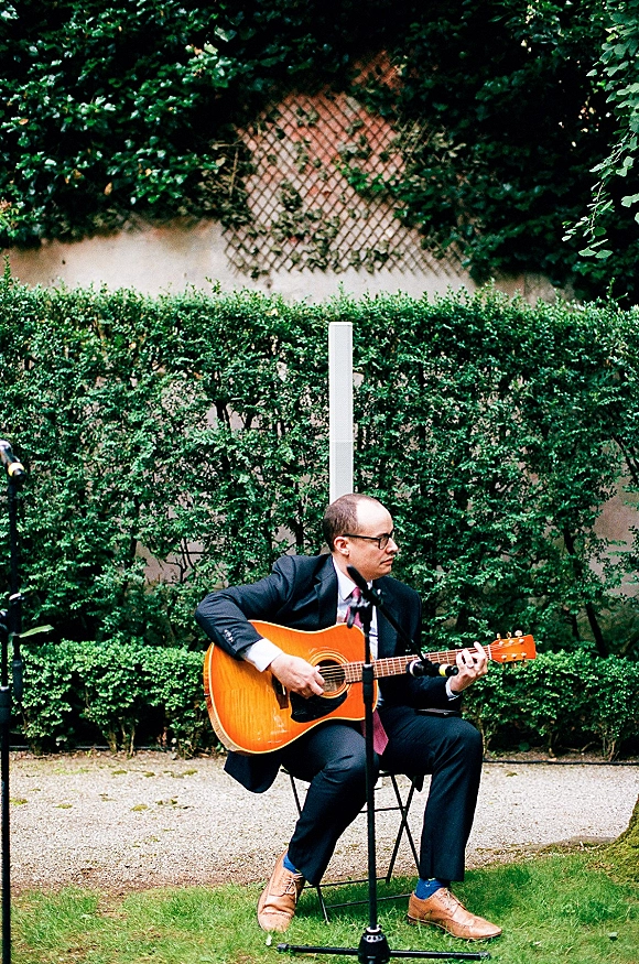 Wedding musician in a suit playing acoustic guitar beside a microphone stand, seated on a folding chair near an ivy wall and lattice fence