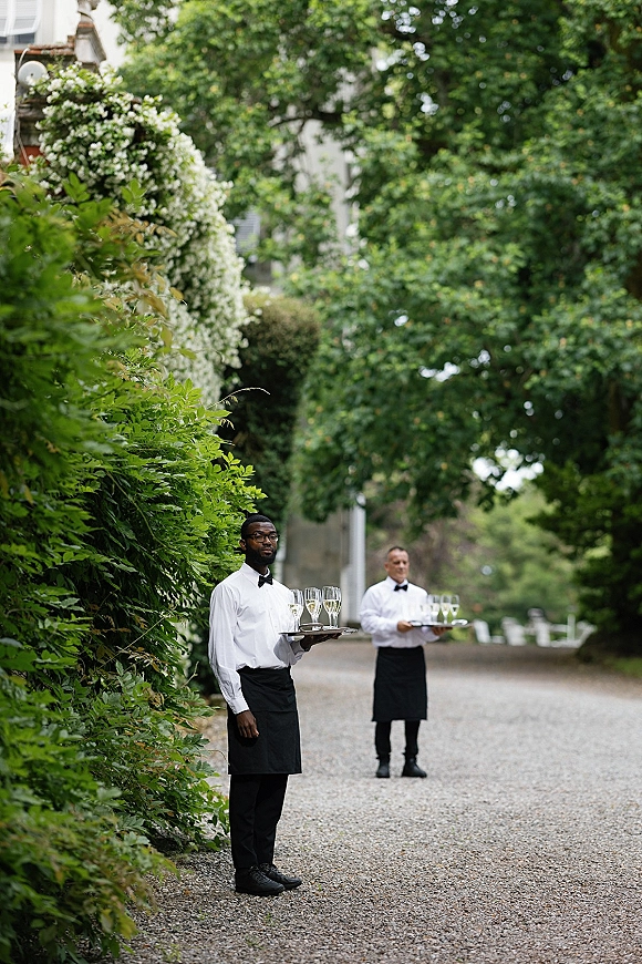 Champagne service with servers in bow ties holding flutes on silver trays along a tree-lined gravel driveway with hedges and vines
