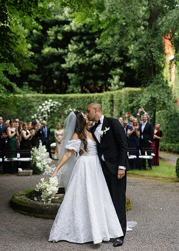 Wedding kiss as bride in off-the-shoulder gown holds a white bouquet, groom in black tuxedo beside garden hedges and guests