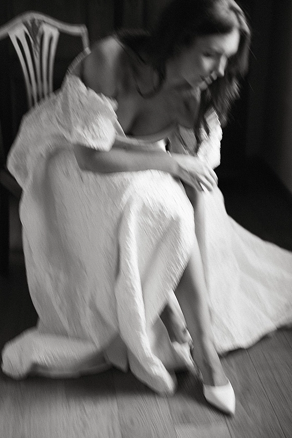 Bride getting ready as she puts on bridal heels, sitting on a chair in an off-the-shoulder wedding dress against a dark wall