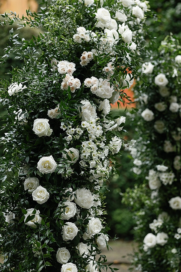 Wedding floral arch with white rose clusters and greenery foliage, arranged on a round frame against softly blurred garden greenery background
