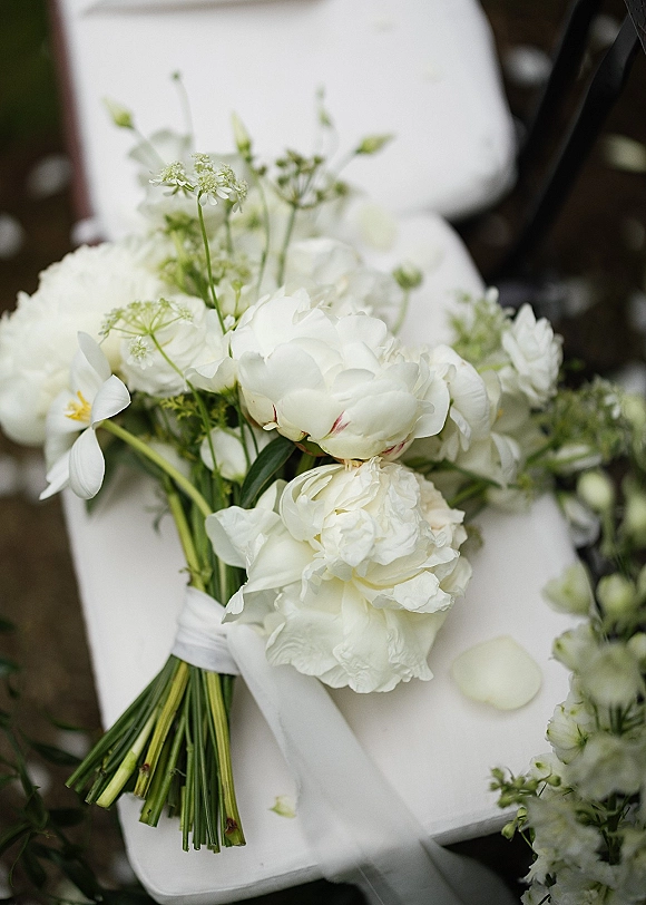 Bridal bouquet with white peony bouquet blooms, cosmos, queen anne’s lace and greenery, ribbon-wrapped, resting on a white chair outdoors