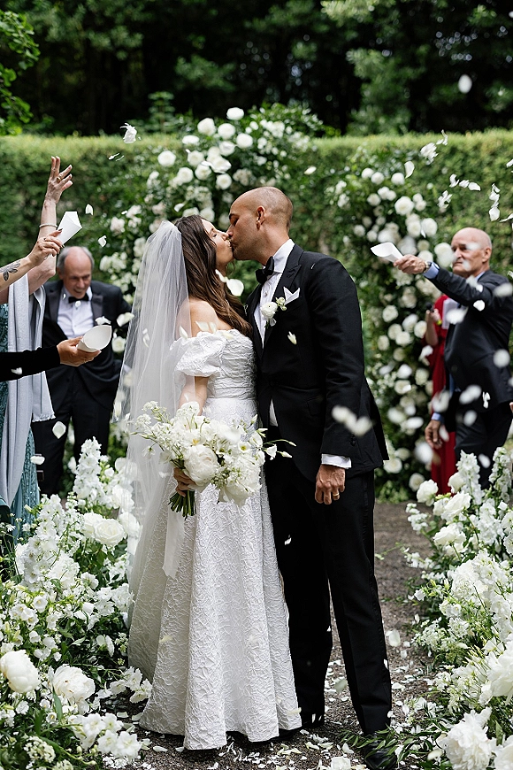 Wedding kiss as newlyweds embrace under a floral arch, bride in lace dress and veil holding bouquet while guests toss petals in garden aisle