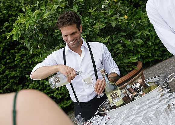 Wedding bartender pouring sparkling wine into a champagne flute, wearing white shirt and suspenders beside an ice tub in a garden setting
